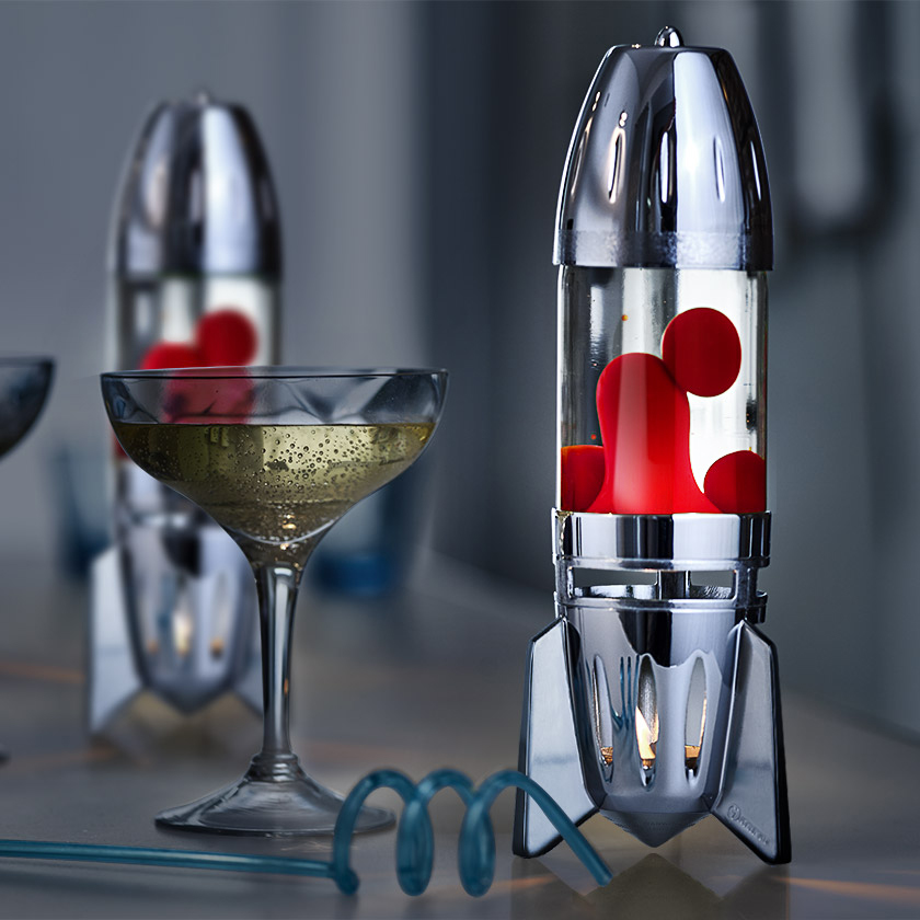 Lampe à lave à bougie argentée en forme de fusée Mathmos Fireflow avec liquide transparent et lave rouge sur une table à côté d'un verre à cocktail