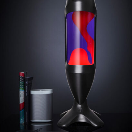 Grande lampe à lave noire Mathmos iO avec liquide violet et lave rouge sur un fond sombre à côté de livres et d'un haut-parleur.