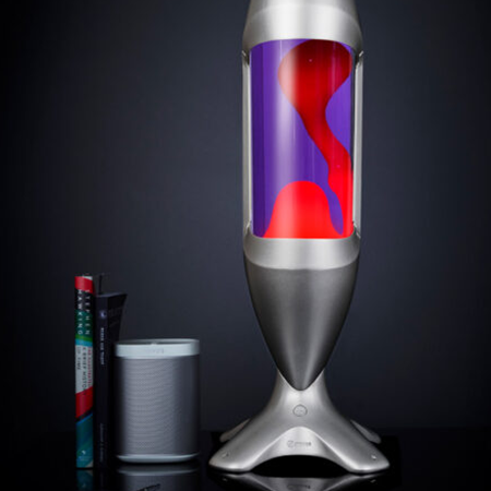 Grande lampe à lave argentée Mathmos iO avec liquide violet et lave rouge sur un fond sombre à côté de livres et d'un haut-parleur.